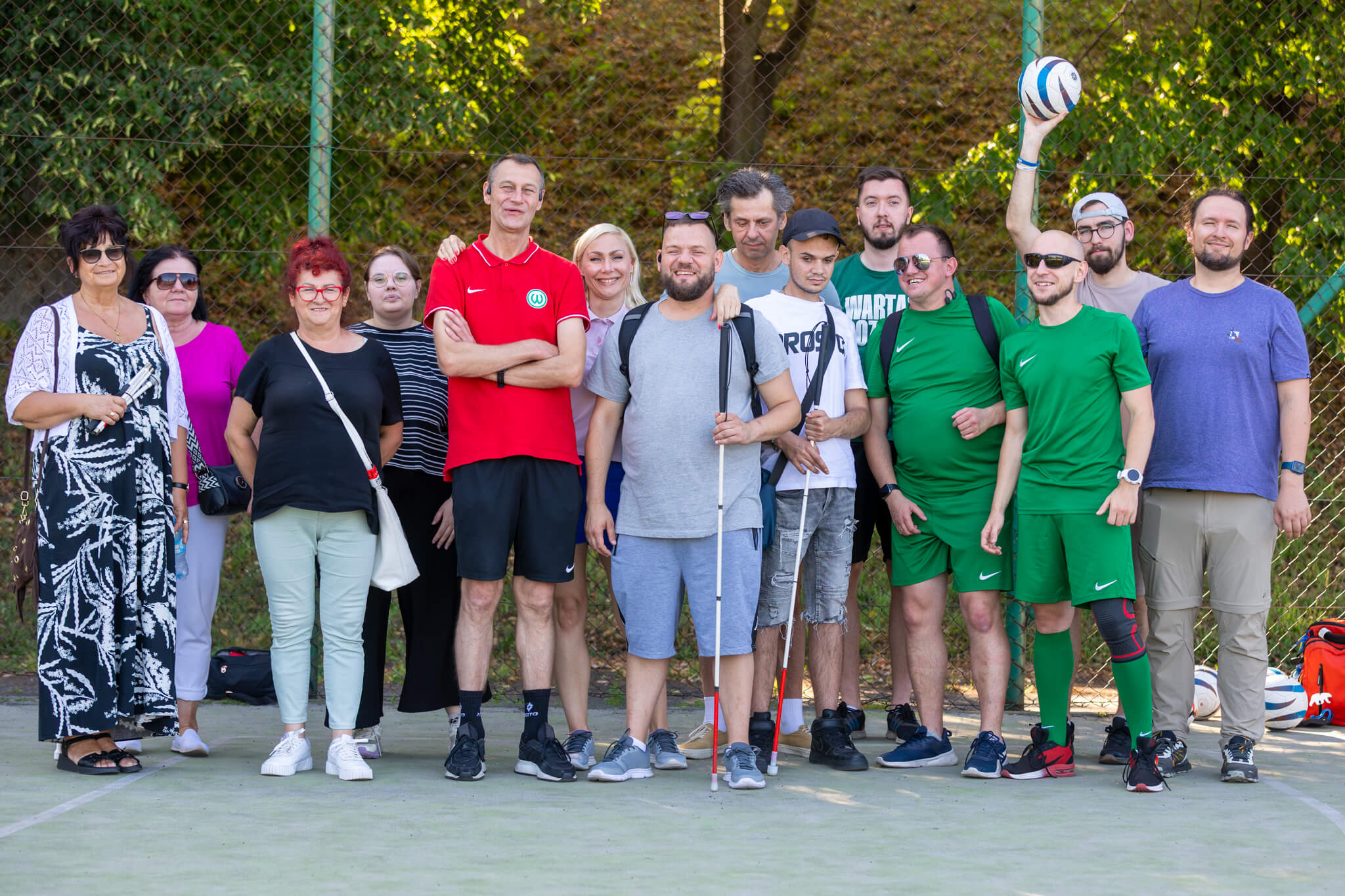 Grupa uczestników i&nbsp;organizatorów pozuje do wspólnego zdjęcia na boisku sportowym. Wśród nich są osoby z&nbsp;białymi laskami oraz zawodnicy ubrani w&nbsp;zielone stroje sportowe. W&nbsp;tle znajduje się ogrodzenie i&nbsp;drzewa, a&nbsp;jedna z&nbsp;osób trzyma nad głową piłkę.