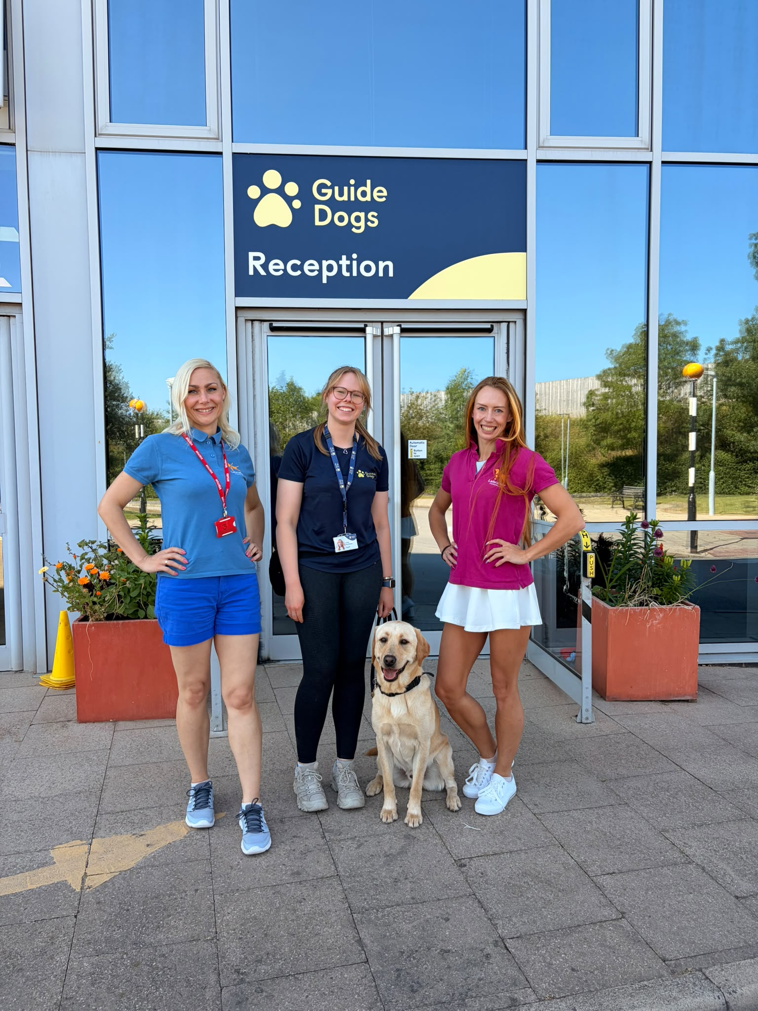 Trzy uśmiechnięte kobiety stoją przed wejściem do ośrodka szkoleniowego Guide Dogs pod tablicą z&nbsp;napisem „Guide Dogs Reception”. Pomiędzy nimi siedzi spokojnie żółty labrador. Wszystkie patrzą w&nbsp;stronę aparatu, a&nbsp;w tle widać przeszkloną fasadę budynku oraz donice z&nbsp;kwiatami. Zdjęcie dokumentuje wizytę w&nbsp;brytyjskim ośrodku szkolenia psów przewodników i&nbsp;wspólne spotkanie z&nbsp;jego pracownikami.