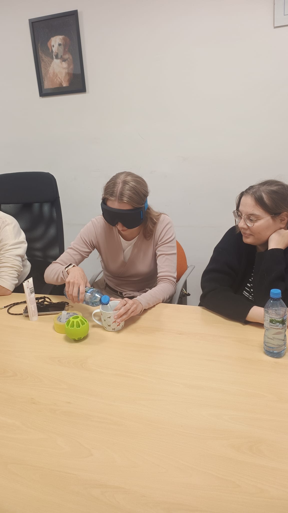 Kobieta w&nbsp;goglach zasłaniających oczy siedzi przy stole i&nbsp;nalewa wodę z&nbsp;butelki do kubka. Obok niej siedzi druga kobieta i&nbsp;obserwuje wykonywane zadanie