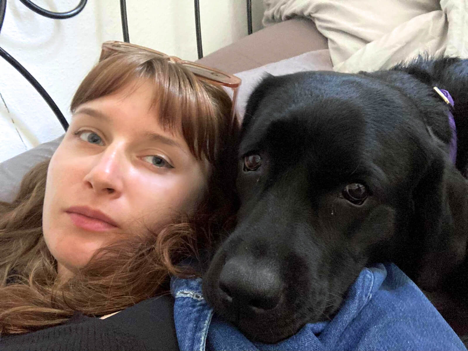 Czarny labrador przytula się do swojej opiekunki podczas wspólnego odpoczynku na łóżku. Pies opiera głowę o&nbsp;jej ramię i&nbsp;spokojnie patrzy przed siebie, podkreślając bliską więź oraz poczucie bezpieczeństwa między nimi.