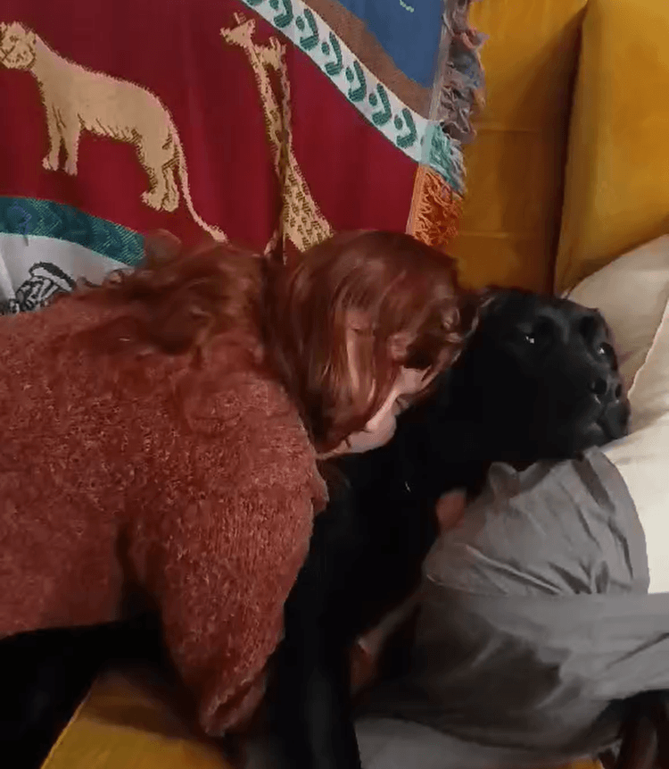 Opiekunka przytula czarnego labradora leżącego na kanapie. Pies spokojnie odpoczywa z&nbsp;głową opartą na poduszce, pozwalając się przytulić. Zdjęcie pokazuje chwilę wyciszenia oraz silną więź i&nbsp;zaufanie między psem a&nbsp;opiekunką.