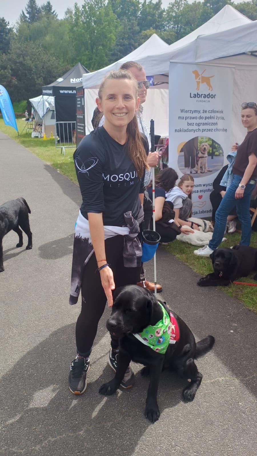 Kobieta stoi z&nbsp;czarnym labradorem w&nbsp;zielonej chustce przed stoiskiem Fundacji Labrador podczas plenerowego wydarzenia. W&nbsp;tle znajdują się namioty wystawców, baner fundacji oraz osoby odwiedzające wydarzenie, a&nbsp;obok odpoczywają inne psy. Zdjęcie przedstawia udział psa asystującego w&nbsp;wydarzeniu publicznym oraz jego przygotowanie do spokojnej pracy wśród ludzi i&nbsp;innych zwierząt.