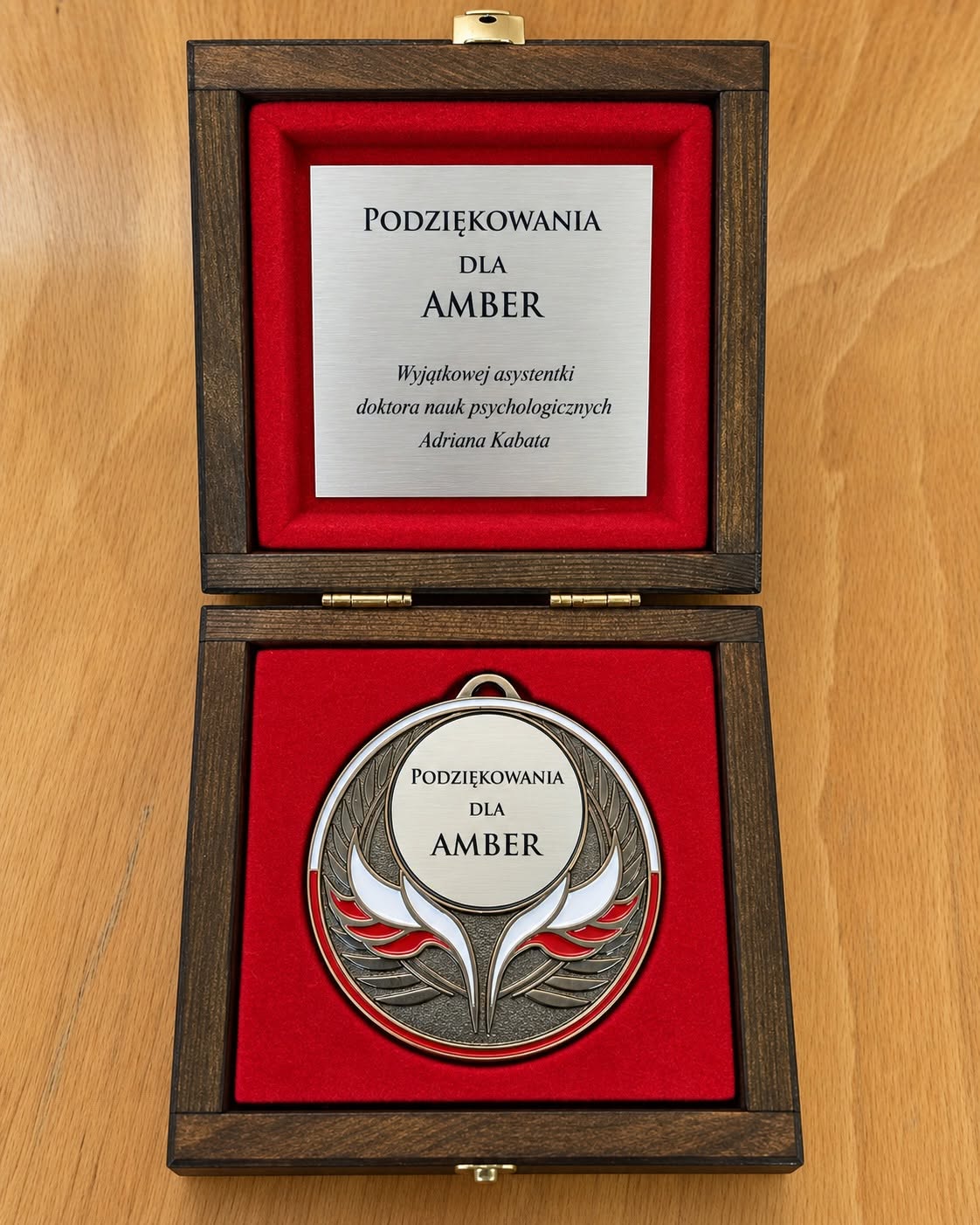 Otwarte drewniane etui wyłożone czerwonym materiałem zawiera okrągły, ozdobny medal oraz metalową tabliczkę z&nbsp;dedykacją. Na tabliczce widnieje napis „Podziękowania dla Amber. Wyjątkowej asystentki doktora nauk psychologicznych Adriana Kabata”. Na medalu również umieszczono podziękowanie dla Amber. Zdjęcie dokumentuje wyróżnienie przyznane labradorce za jej rolę psa asystującego, podkreślając znaczenie jej codziennej pomocy oraz obecności w&nbsp;życiu zawodowym i&nbsp;akademickim opiekuna.