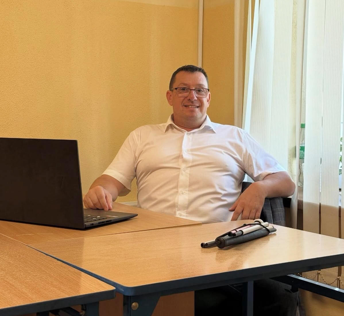 Na zdjęciu widoczny jest mężczyzna siedzący przy biurku w&nbsp;jasnym, biurowym pomieszczeniu. Ma na sobie białą koszulę z&nbsp;krótkim rękawem i&nbsp;okulary. Siedzi w&nbsp;swobodnej, lekko odchylonej pozycji, z&nbsp;jedną ręką opartą o&nbsp;blat, a&nbsp;drugą na oparciu krzesła.  Na biurku stoi otwarty laptop, a&nbsp;obok niego leżą złożona biała laska dla osób niewidomych oraz jej elementy. W&nbsp;tle widać jasną ścianę oraz okno z&nbsp;pionowymi żaluzjami, przez które wpada naturalne światło.